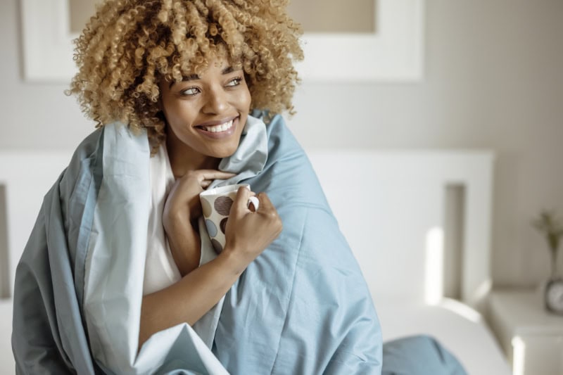 5 Reasons to Schedule a Fall Furnace Clean and Check. Pretty Woman Snuggled Under Duvet With Cup of Coffee or Tea in Bed at Home Bedroom. Morning, Leisure and People Concept.