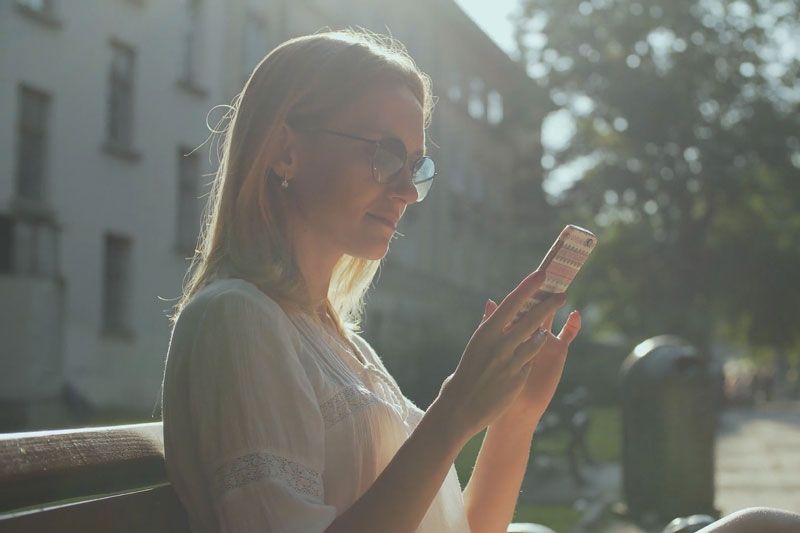 Video - Are Wi-Fi Thermostats Cost-Effective?. Image shows a woman with blonde hair wearing sunglasses and sitting on a bench outside and looking at her cell phone.
